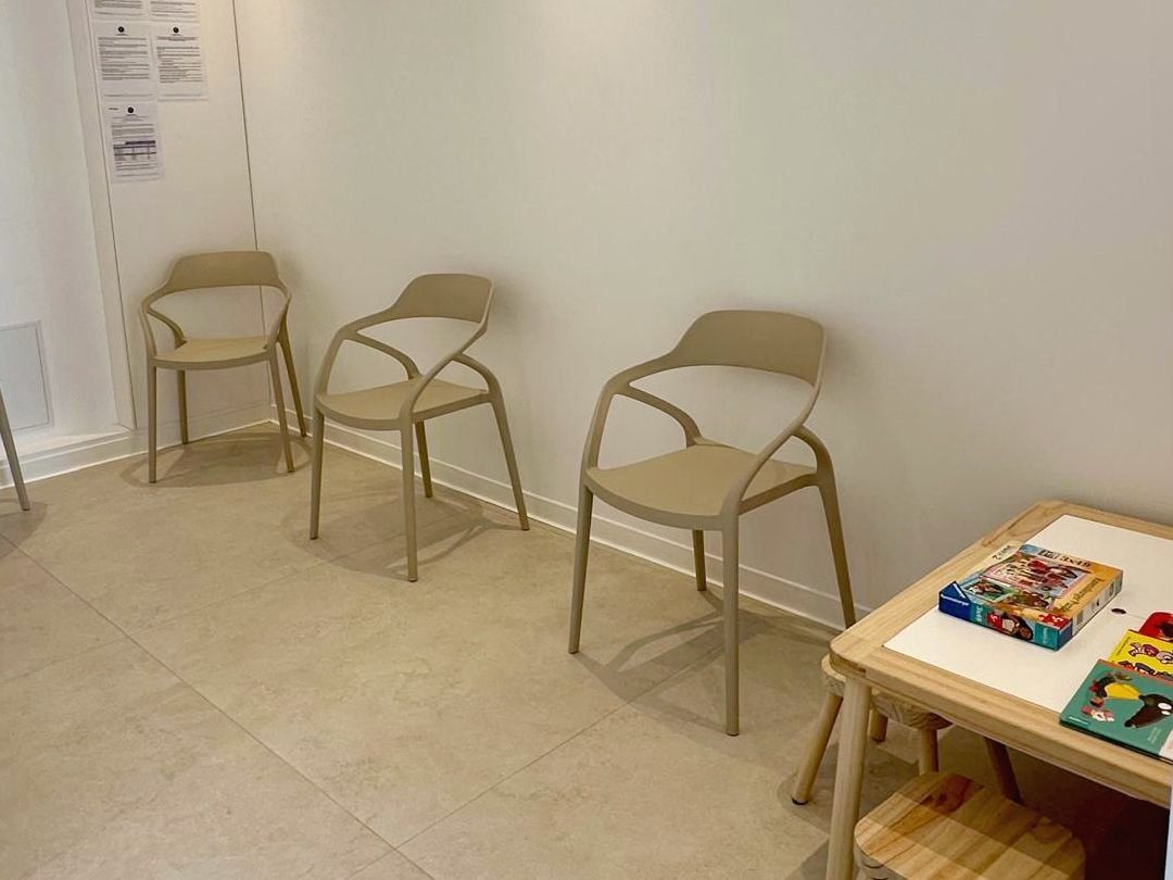 Salle d’attente claire : chaises beiges alignées le long d’un mur blanc et, au premier plan, le coin jeux des enfants avec une petite table en bois, deux tabourets, des livres et un jeu.