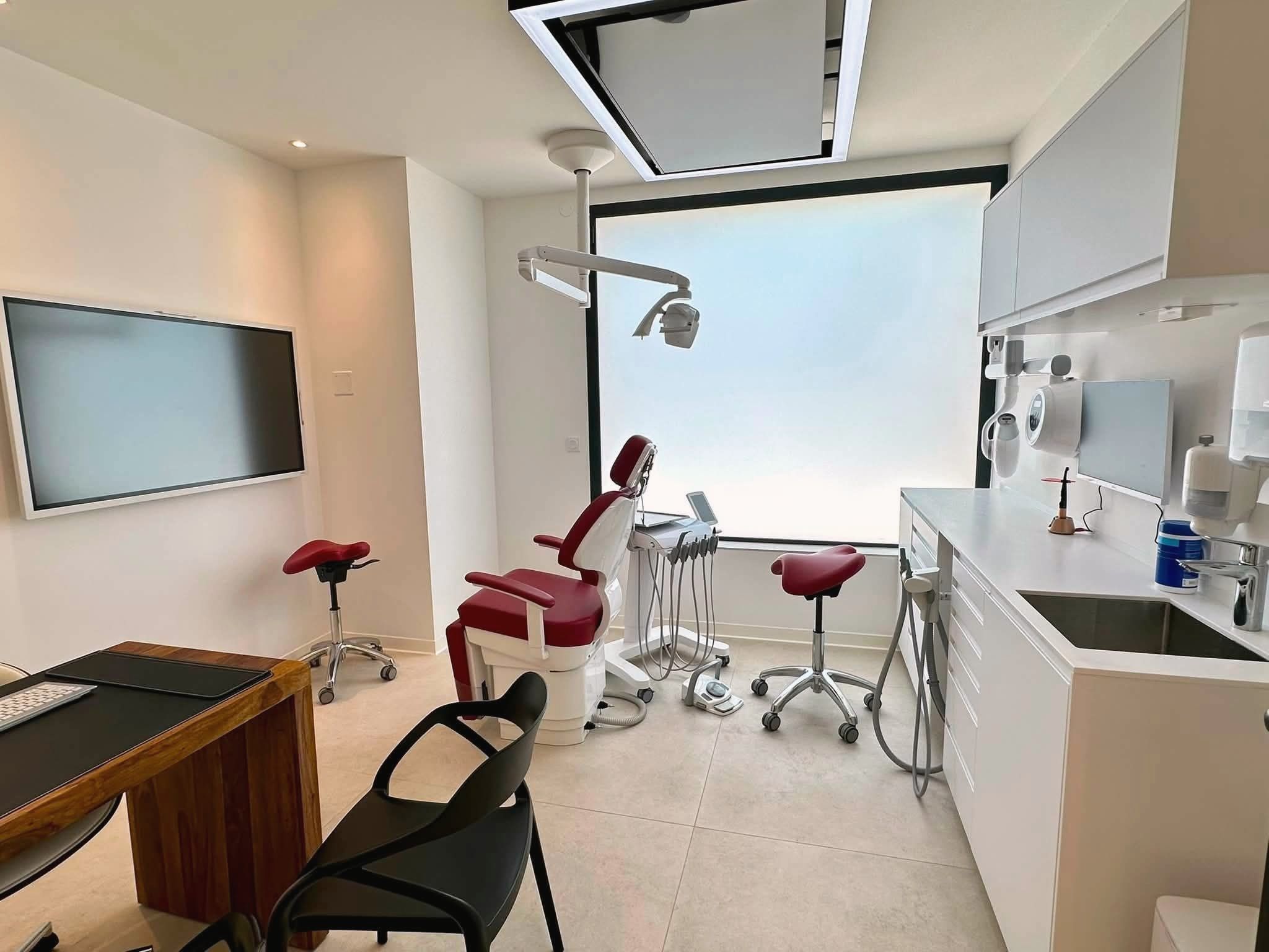 Salle de soins principale : fauteuil et tabourets rouge grenat devant une grande baie en verre dépoli, écran au plafond, grand écran tactile au mur et bureau en bois au premier plan.