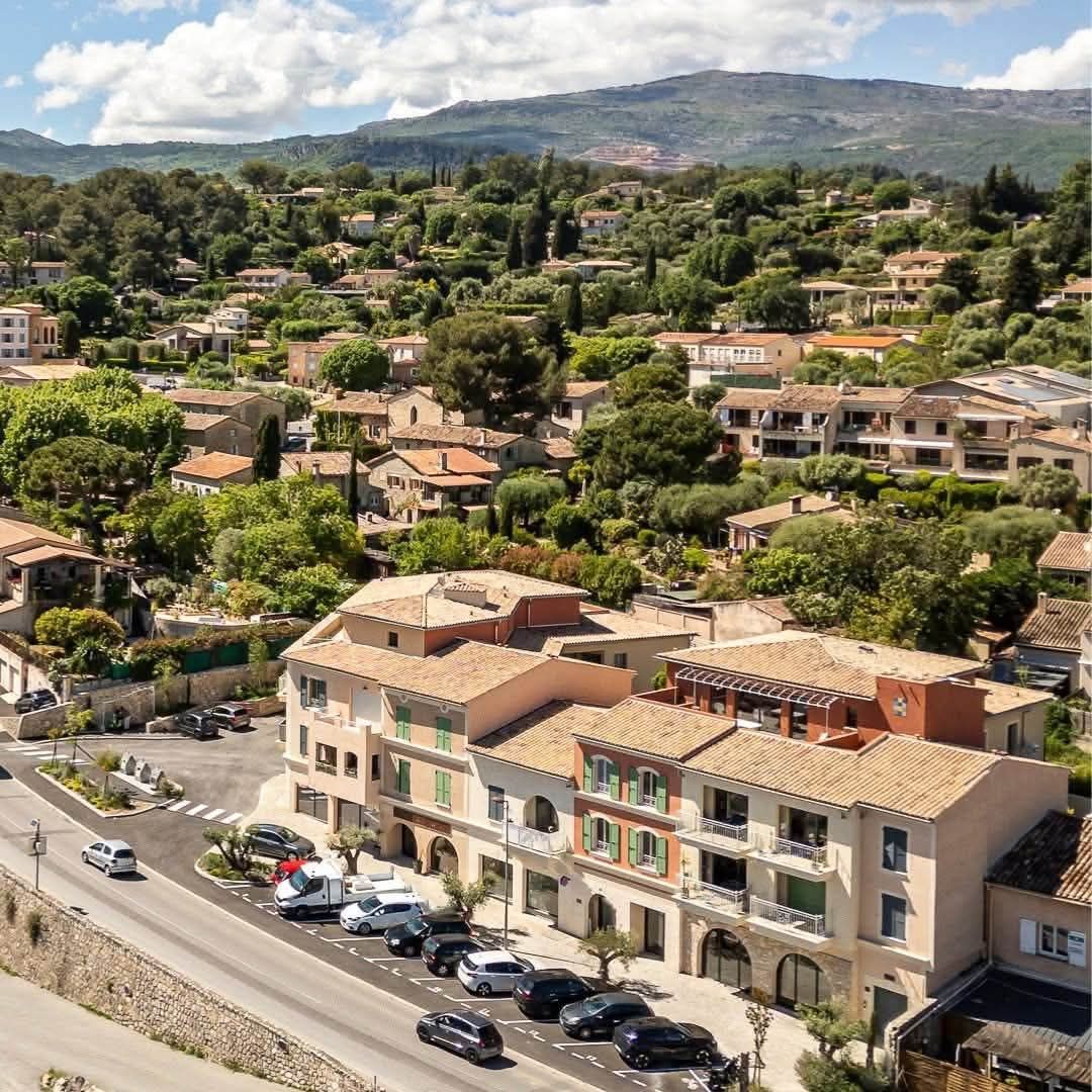 Vue aérienne du Rouret : au premier plan, l’immeuble neuf à arcades de l’avenue de Nice, livré en 2025, où se trouve le cabinet, et les places de stationnement devant ; derrière, le village dans la verdure et les Préalpes.